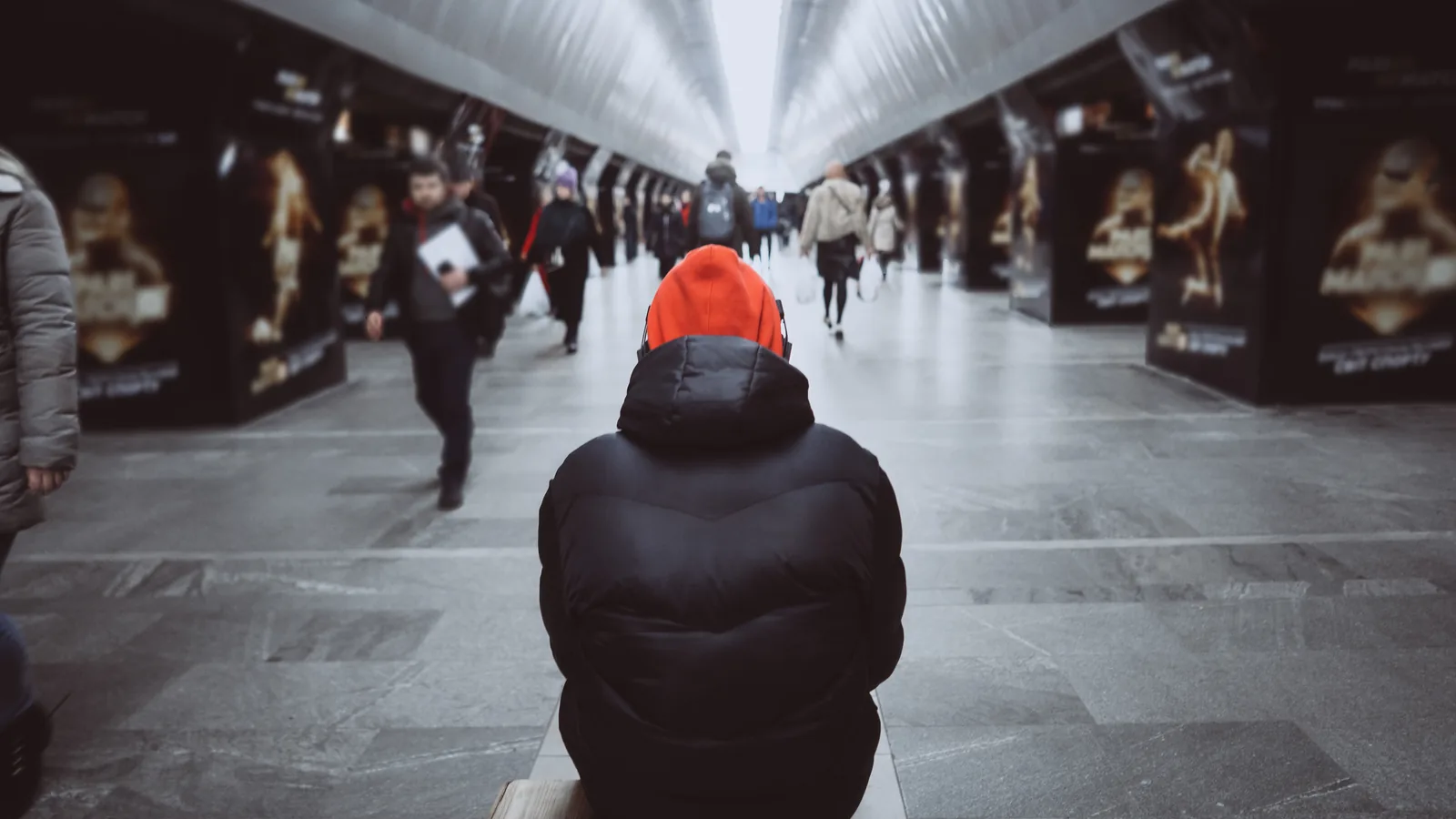 Man in a red hoodie sitting still in a busy metro corridor while commuters walk around him.
